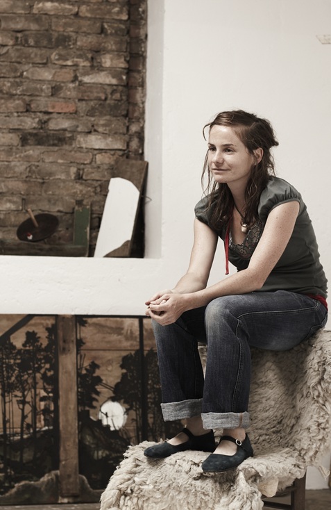 It shows the artist in her studio sitting in a chair with her hands together.