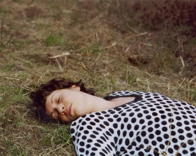 The artist is shown lying on the grass, wearing a white shirt with black polka dots.